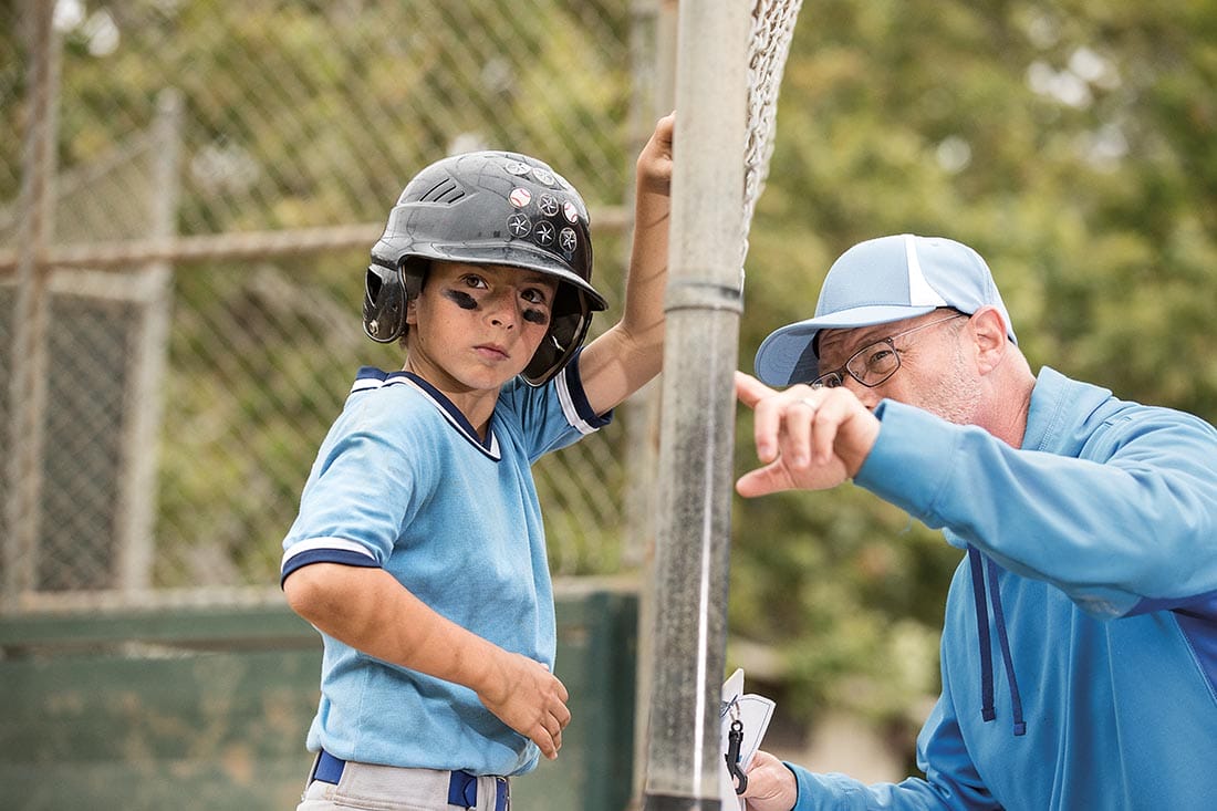 A young boy wearing a blue baseball uniform talking to a coach through a chain-link fence