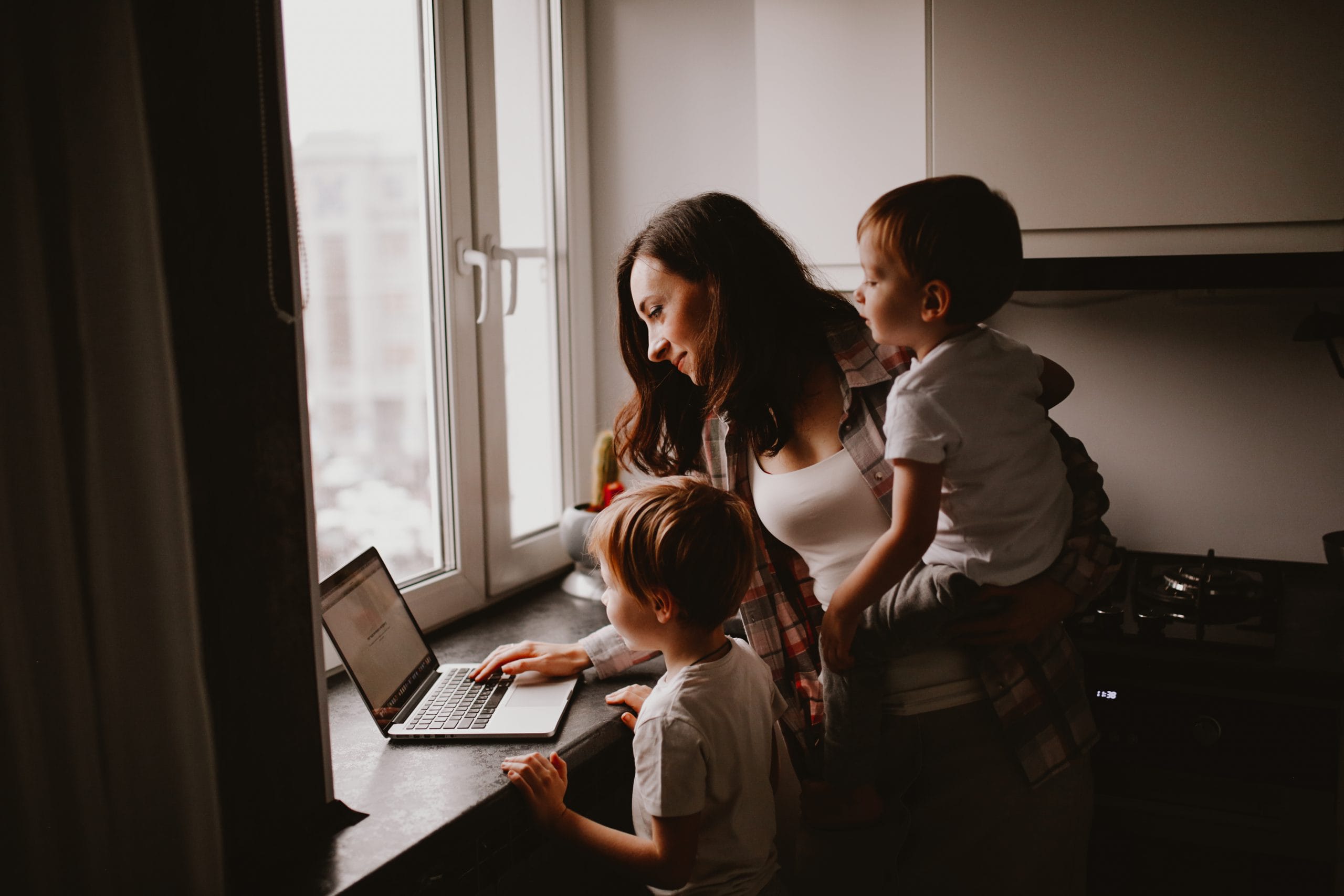 Busy cute woman mom with two children works on a laptop in the kitchen and talks on the phone, mom turns on the kids cartoon. Family concept and freelance work, authentic lifestyle and toning