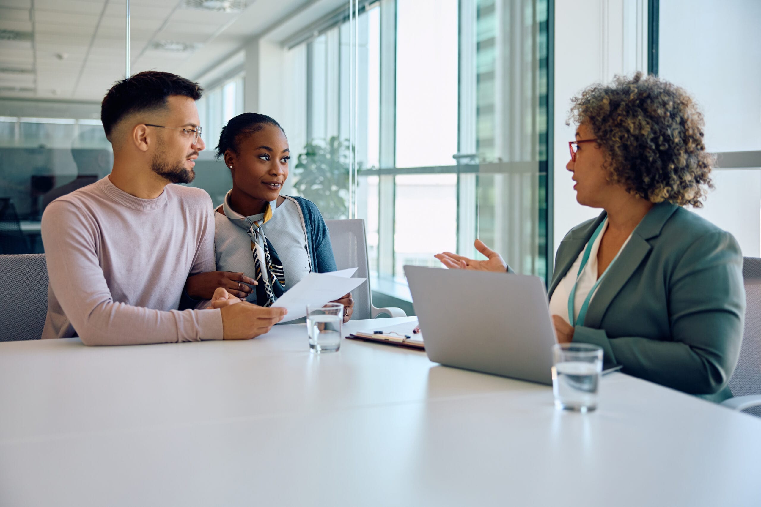 Insurance producer speaking with a laptop open in front of her as she sits with a multiracial couple reviewing a piece of paper in an office.